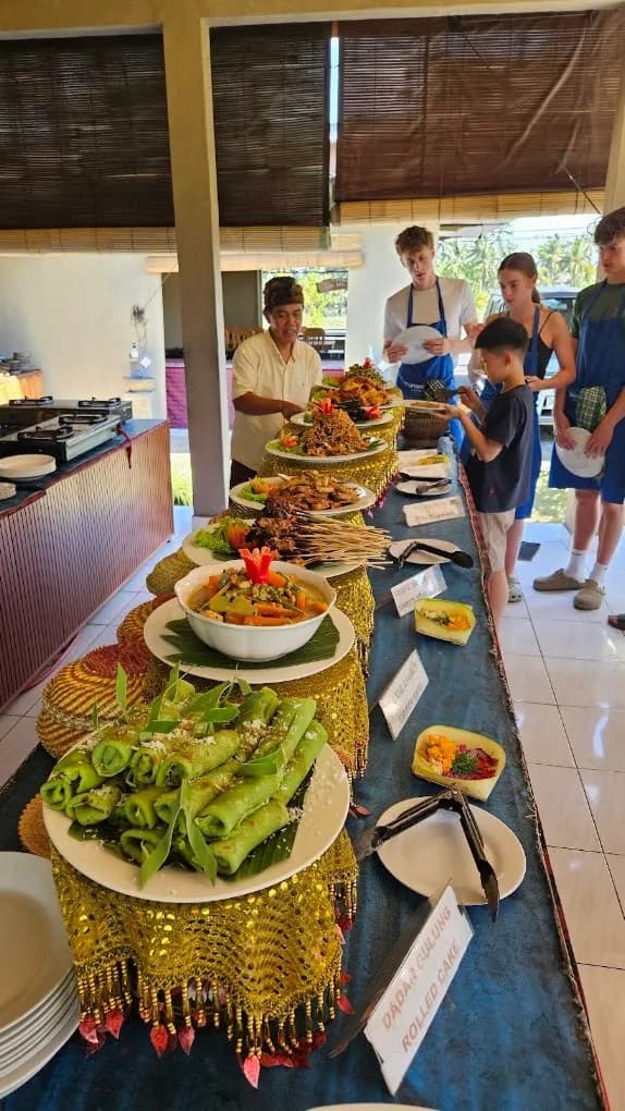 Guests with plates at the Petulu class buffet, dadar gulung labeled at the front of the line, on a food day near Ubud