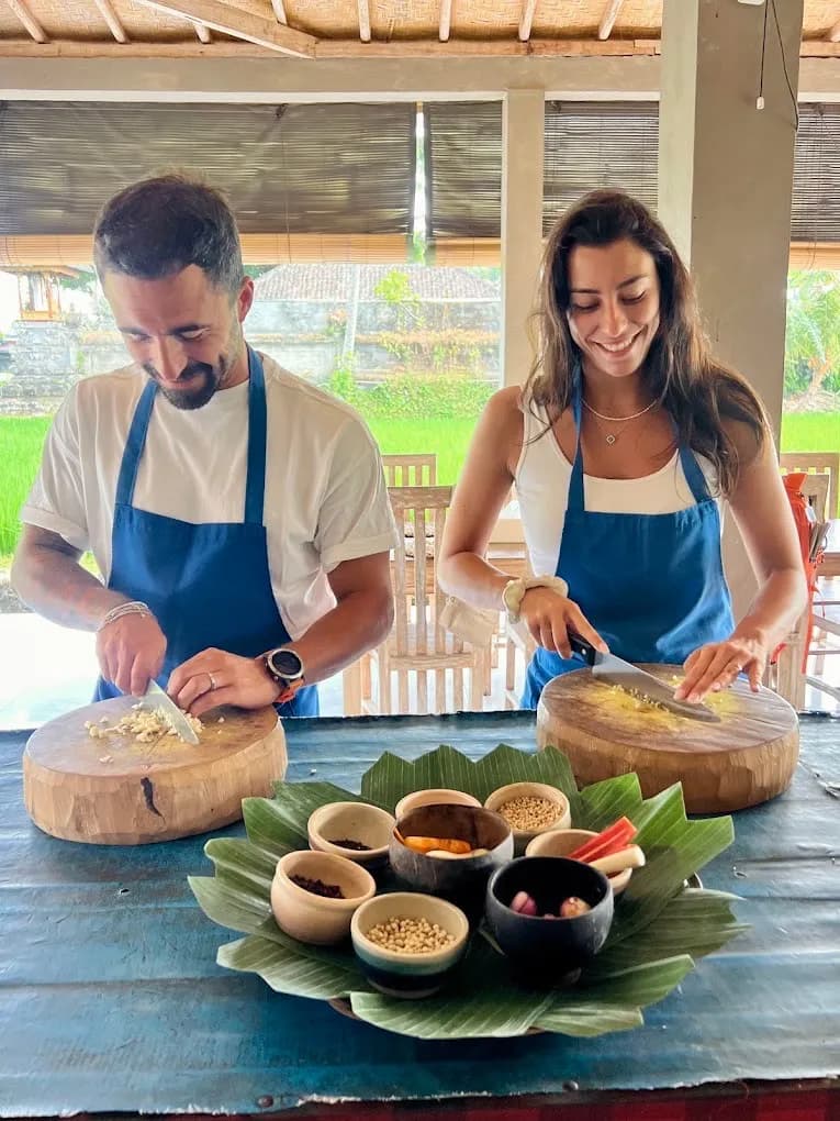 Two guests in blue aprons chopping garlic on wooden blocks beside a banana-leaf tray of spices, rice field behind, in Petulu, on a food day near Ubud